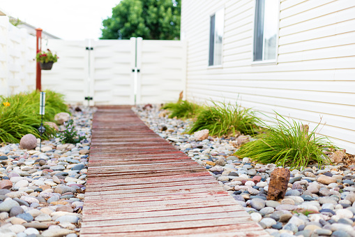 In Western Colorado Side yard of Residential Home with wooden red paint worn walkway lined with decorative gray river rock and flowering bushes surrounded by edge of house with two windows and white fence with gate at far end and a lone flower pot hanging from a post (Shot with Canon 5DS 50.6mp photos professionally retouched - Lightroom / Photoshop - original size 5792 x 8688 downsampled as needed for clarity and select focus used for dramatic effect)