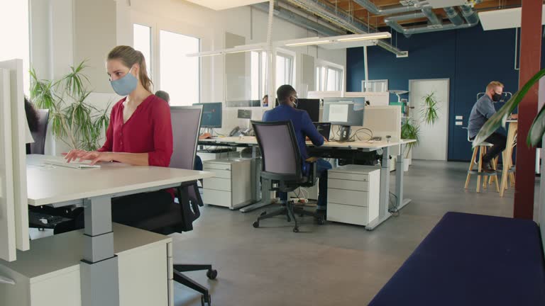 Business people wearing face mask working in office. Business people back to work sitting at desk with protection guard between them post corona virus lockdown.