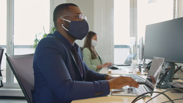 Businessman with protective face mask working at his desk. Business people return back to work after pandemic lockdown sitting at desk with protection guard between them.
