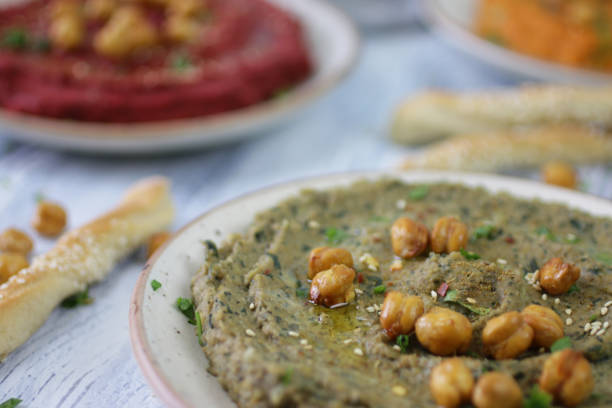 Stock photo showing bowls containing sundried tomato and basil hummus, healthy eating vegan appetiser, Greek cuisine food (hommus / houmous) made from a base of chickpeas, olive oil, tahini paste and garlic surrounded by home-made breadstick twists topped with sesame seeds. imagen de cuencos que contienen albahaca y hummus de tomate seco rodeado de garbanzos asados secos y torceros caseros de palillos cubiertos con semillas de sésamo sobre fondo de madera encalado, cocina griega, se centran en el primer plano