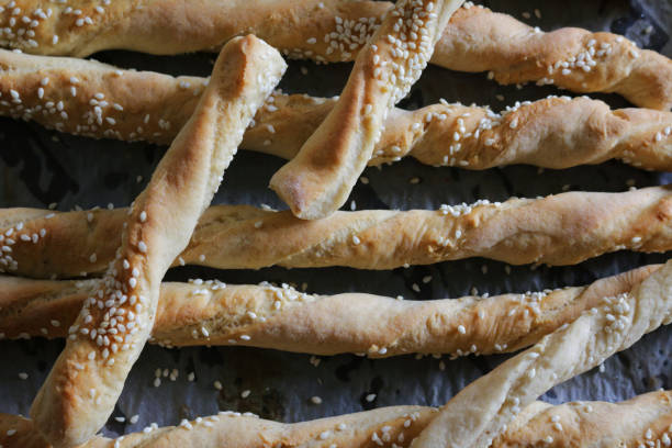 Stock photo showing close-up, elevated view of homemade white breadstick twists on greaseproof paper lined baking tray, ready to eat. imagen de primer plano de los giros de barra de pan horneados y caseros cubierto con semillas de sésamo en bandeja de hornear forrada con papel / pergamino a prueba de grasa, dorado, fresco del horno, giros de pan crujiente, vista elevada