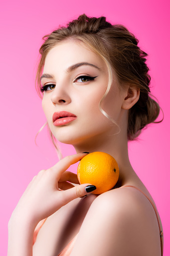 elegant beautiful blonde woman holding ripe orange on shoulder isolated on pink