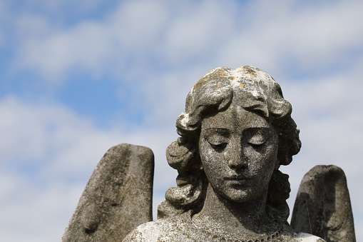 A dark stone angel's solemn regard in a cemetery (Mitcham, Surrey, UK), with a background of blue sky and summer clouds.