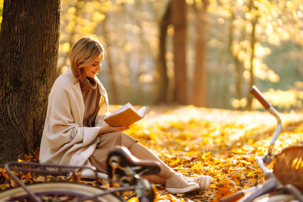 hermosa joven sentada en un otoño caído sale en un parque, leyendo un libro. - autumn women park forest fotografías e imágenes de stock