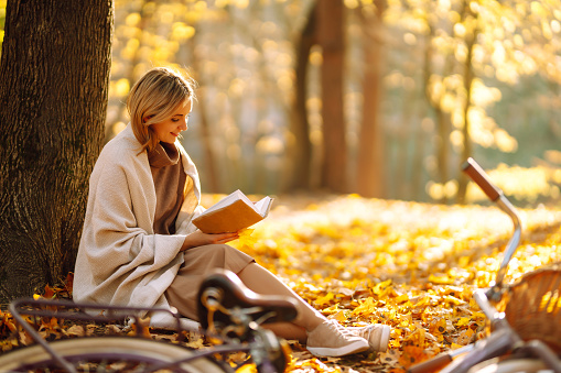 Beautiful young woman sitting on a fallen autumn leaves in a park, reading a book.