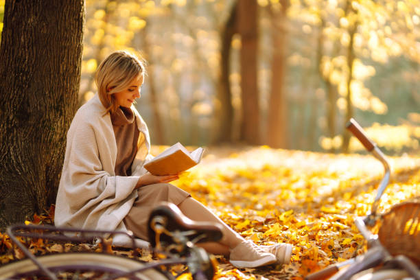 beautiful young woman sitting on a fallen autumn leaves in a park, reading a book. - autumn women park forest imagens e fotografias de stock