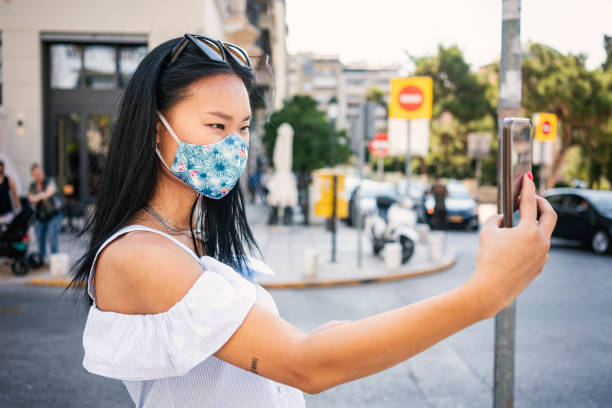 istock Young woman wearing protective face mask walking through city, using smart phone 1264462481