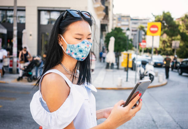 istock Young woman wearing protective face mask walking through city, using smart phone 1264462543