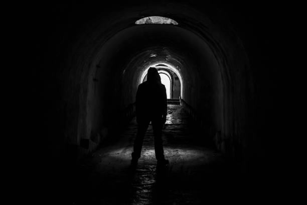 istock A lonely hooded man walks out into the light from a dark, abandoned underground corridor. 1264465624