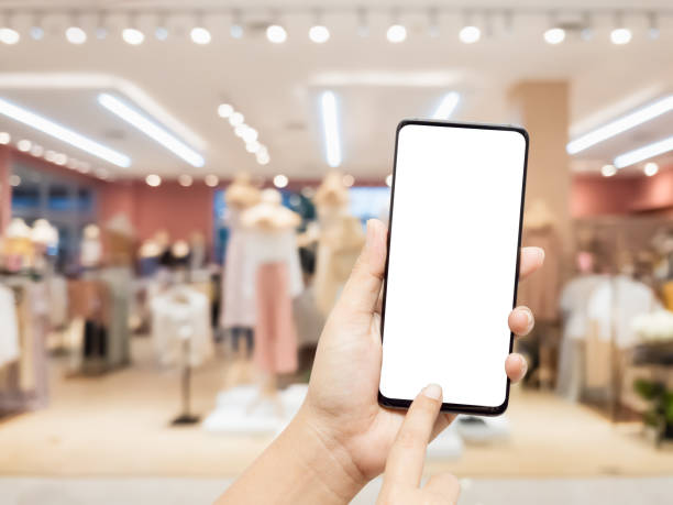 istock Mockup blank white screen mobile phone. Hand holding and pointing to smartphone wirh blurred female clothing store background for your text or advertisement artwork. 1264469502