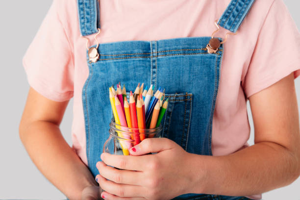 A nine year old student is posing holding a pot full of coloring pencils (face not seen). She is wearing jeans overall. She is in a studio. She is in Quebec, Canada. She is getting ready to go back to school.