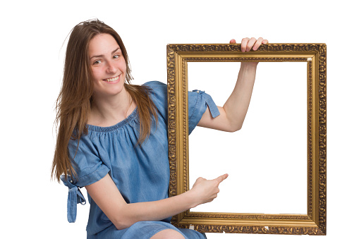A young girl with long hair in a blue dress. Isolated on a white background, holding a picture frame in his hands, pointing at it with his finger. copyspace. with a happy, smiling expression on his face