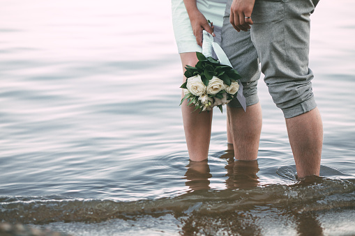 The view of the newlyweds legs standing on the beach. The view of the wedding bouquet of white roses and cotton