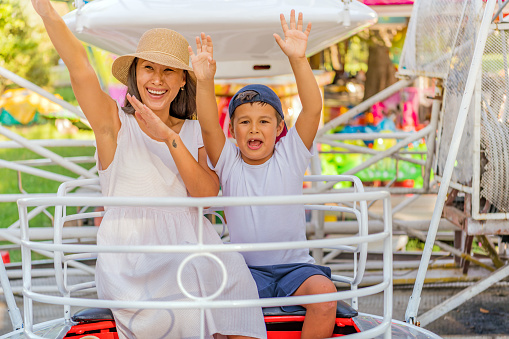 A photo of a mother smiling with her young seven-year-old son while riding a small roller-coaster at an amusement park during a summer day. Different expressions cross the faces of these three fun riders. Beautiful Woman of Asian Ethnicity is Having a Lot of Fun Riding on Ferris Wheel with her Cute Little Son. Happy Mother and Son are Enjoying in Amusement Park on a Rollercoaster on a Warm Sunny Day.