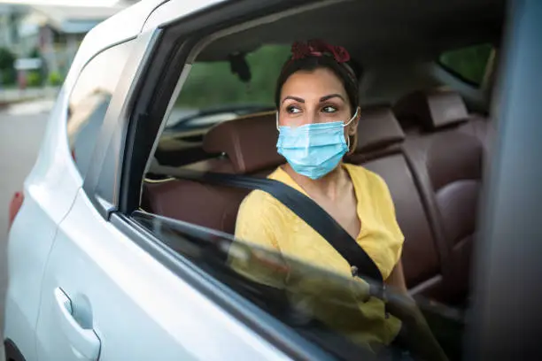 Woman with protective face mask, sitting on back seat of car, looking through the window and enjoying a ride, ride sharing concept
