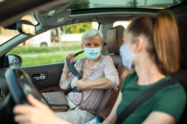 Smiling women with protective face masks in ride sharing car, senior woman fastening seat belt