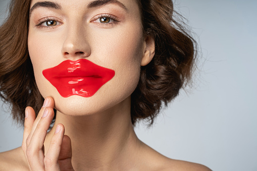 Close up of attractive lady with red lip mask pad on her lips standing against light blue background