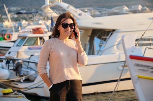 Portrait of mature beautiful woman walking in sea pier talking on mobile phone smartphone, landscape mountains sunset at sea yacht club background