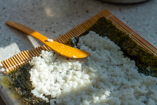 Preparing homemade Sushi.  This shows a sushi bamboo mat with sushi nori seaweed wrapper and sushi rice being spread using spatulas.