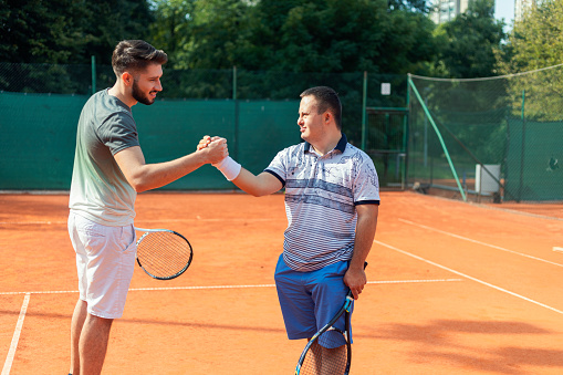 Young man with Down syndrome playing tennis with his friend on clay tennis court