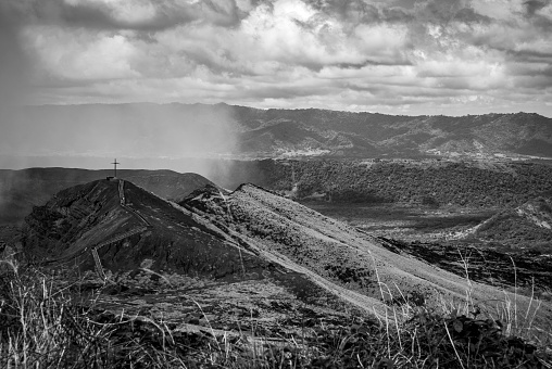 Black and white photograph of the smoking volcanic crater at Masaya Volcano National Park in Nicaragua. A path leads to a cross on the crater's rim.