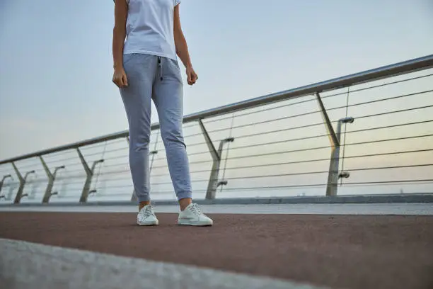 Cropped photo of a fit tanned Caucasian lady in activewear standing on a pedestrian bridge