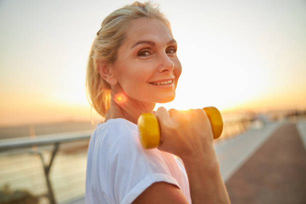 Sporty lady standing on a bridge during the training Portrait of a good-looking cheerful middle-aged athletic woman with a dumbbell posing for the camera healthy lifestyle women outdoors athlete stock pictures, royalty-free photos & images