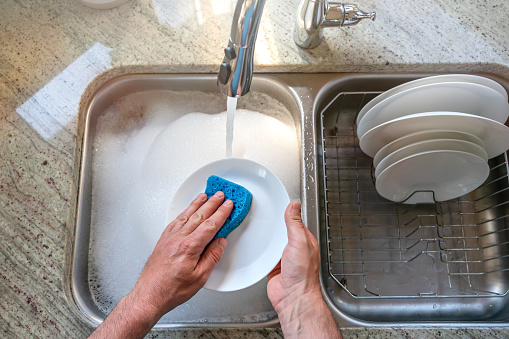 A man washes a white plate with a blue foam sponge in the kitchen, top view. Dishwashing, home routine, cleaning ups the plates. Close up man's hands over the kitchen sink with running water.