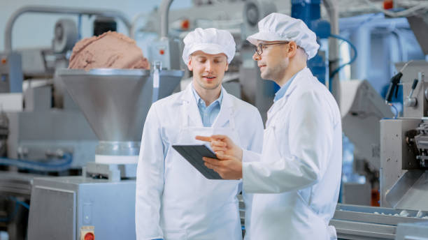 istock Two Young Food Factory Employees Discuss Work-Related Matters. Male Technician or Quality Manager Uses a Tablet Computer for Work. They Wear White Sanitary Hat and Work Robes. 1264531402