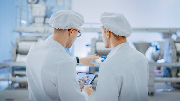 istock Two Young Food Factory Employees Discuss Work-Related Matters. Male Technician or Quality Manager Uses a Tablet Computer for Work. They Wear White Sanitary Hat and Work Robes. 1264531411