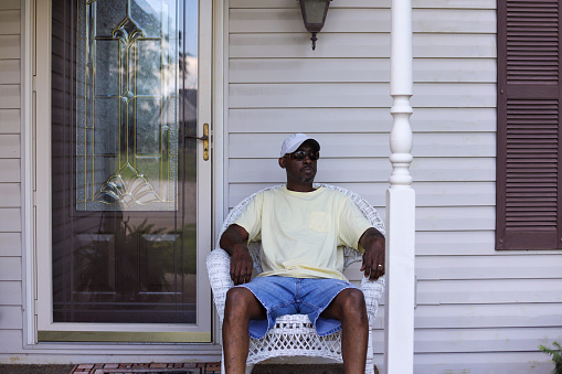 A portrait of an African-American man sitting alone in a wicker chair on a porch and feeling sad and lonely