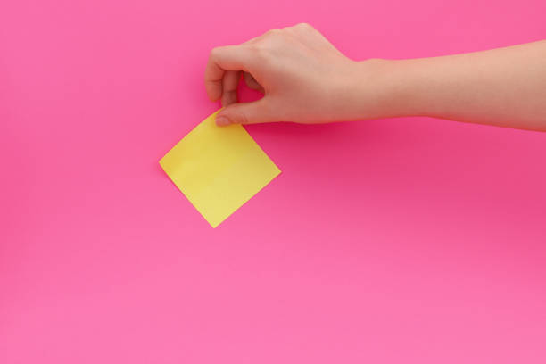 Woman's hand glues yellow sticker or sticky post-it notes on pink background. stock photo
