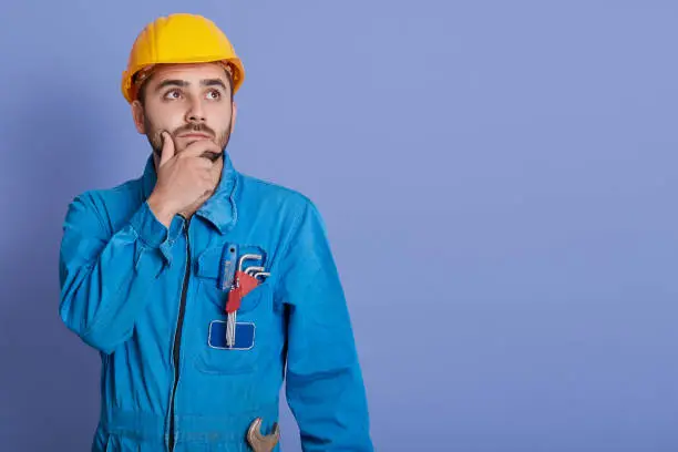 Photo of Pensive guy wearing yellow helmet and blue overalls, bared boy with wrench, looking aside with thoughtful facial expression. Copy space for advertisement.
