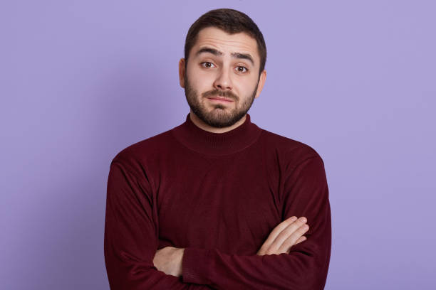 istock Thoughtful young man with skeptic, doubtful, distrustful look posing against lilac background with folded hands, wearing burgundy sweater, bearded puzzled male. 1264542632