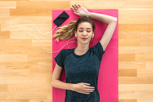 Top view of beautiful blonde sportswoman smiling and lying on yoga mat on wooden floor, listening music with earphones using smartphone with eyes closed.