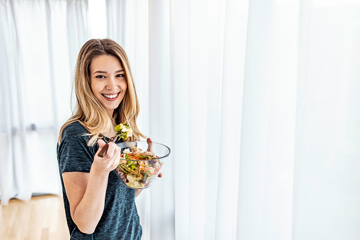 Portrait of a young happy playful girl eating fresh salad from a bowl, looking at camera while standing next to big bright window at home.
