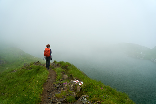 Woman with backpack hiking on a trail by a glacial lake in the mist