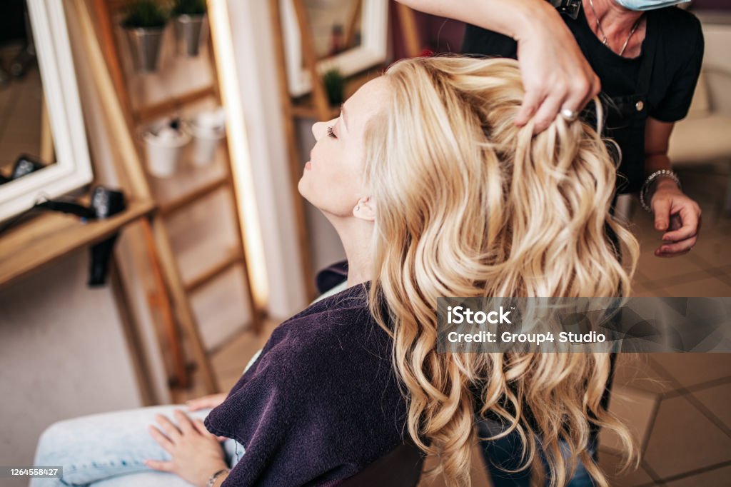 Woman with protective mask receiving treatment in hair salon Professional hairdresser making a beautiful hairstyle of young adult woman after dyeing hair and making highlights in hair salon. Modern lifestyle concept. Hair Salon Stock Photo