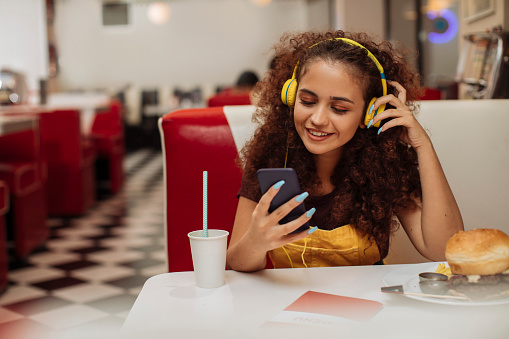 Young curly woman with headphones eating a burger in an American style diner