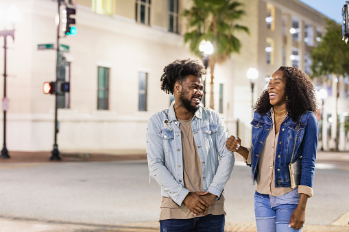 A young African-American couple hanging out in the city, talking  and smiling as they standing on a street corner. It is evening, with the street lights illuminated.