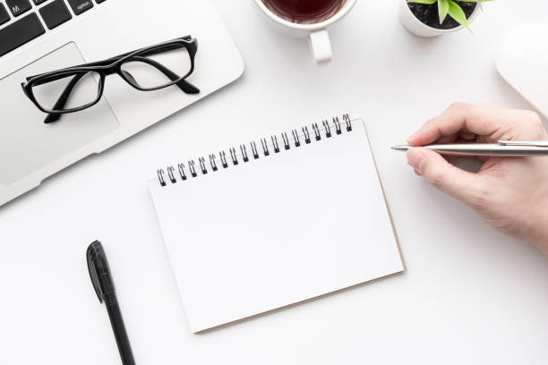 istock Man hand is going to write something on blank notebook page over white office desk table. Top view with copy space, flat lay. 1264593596