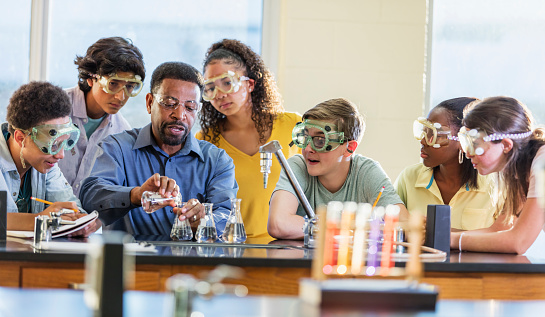 A mature African-American man in his 50s teaching a multi-ethnic group of teenagers in a high school science class. He is performing a chemistry experiment with beakers and test tubes.  The students are watching as he pours chemicals from one beaker into another.