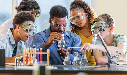 A mature African-American man in his 50s teaching a multi-ethnic group of teenagers in a high school science class. He is demonstrating a chemistry experiment with beakers and test tubes.  Everyone is staring at the beaker he is holding aloft.