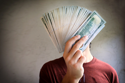 close up image of young male hands holding pile of money and covering his face