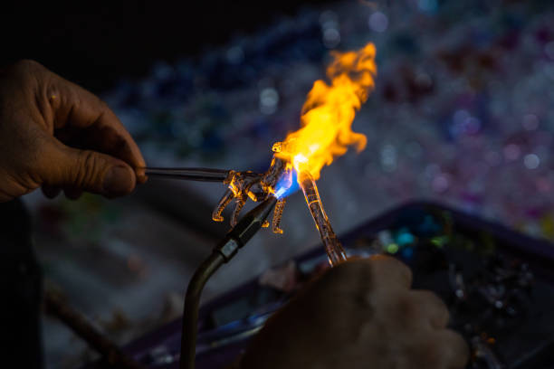 Process of glass blowing melting glass into small figurine using torch with orange flames. Part of human hand. Blurred bokey background.