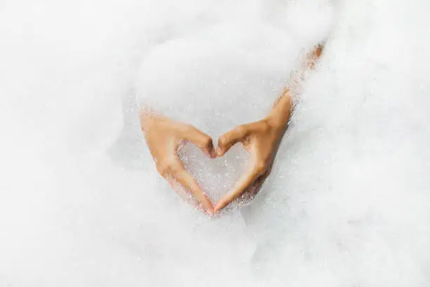 Photo of Woman hands in heart shape in bath full of foam bubbles close-up. Spa beauty treatment. Love symbol