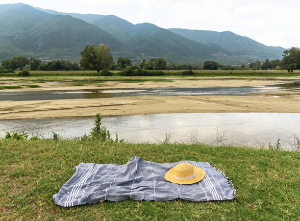 Straw hat at striped blanket at green grass with river and mountain at background