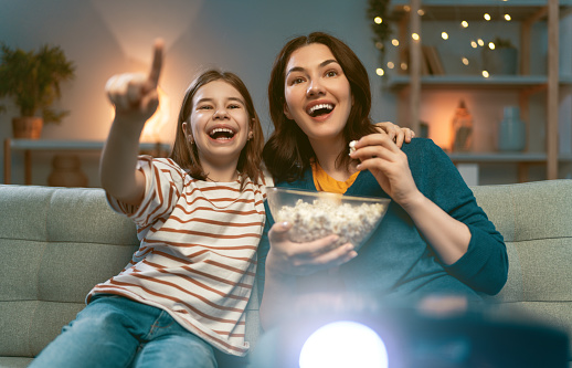 Happy family watching projector, TV, movies with popcorn in the evening at home. Mother and daughter spending time together.