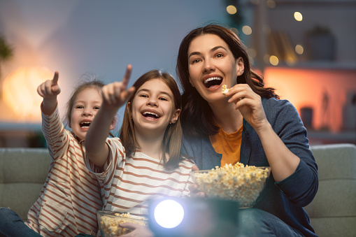 Happy family watching projector, TV, movies with popcorn in the evening at home. Mother and daughters spending time together.