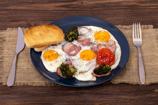 istock Savoury breakfast with fried eggs, bacon, tomato, broccoli and toasts in a dark plate that is on burlap, with fork and knife, angle view 1264642125